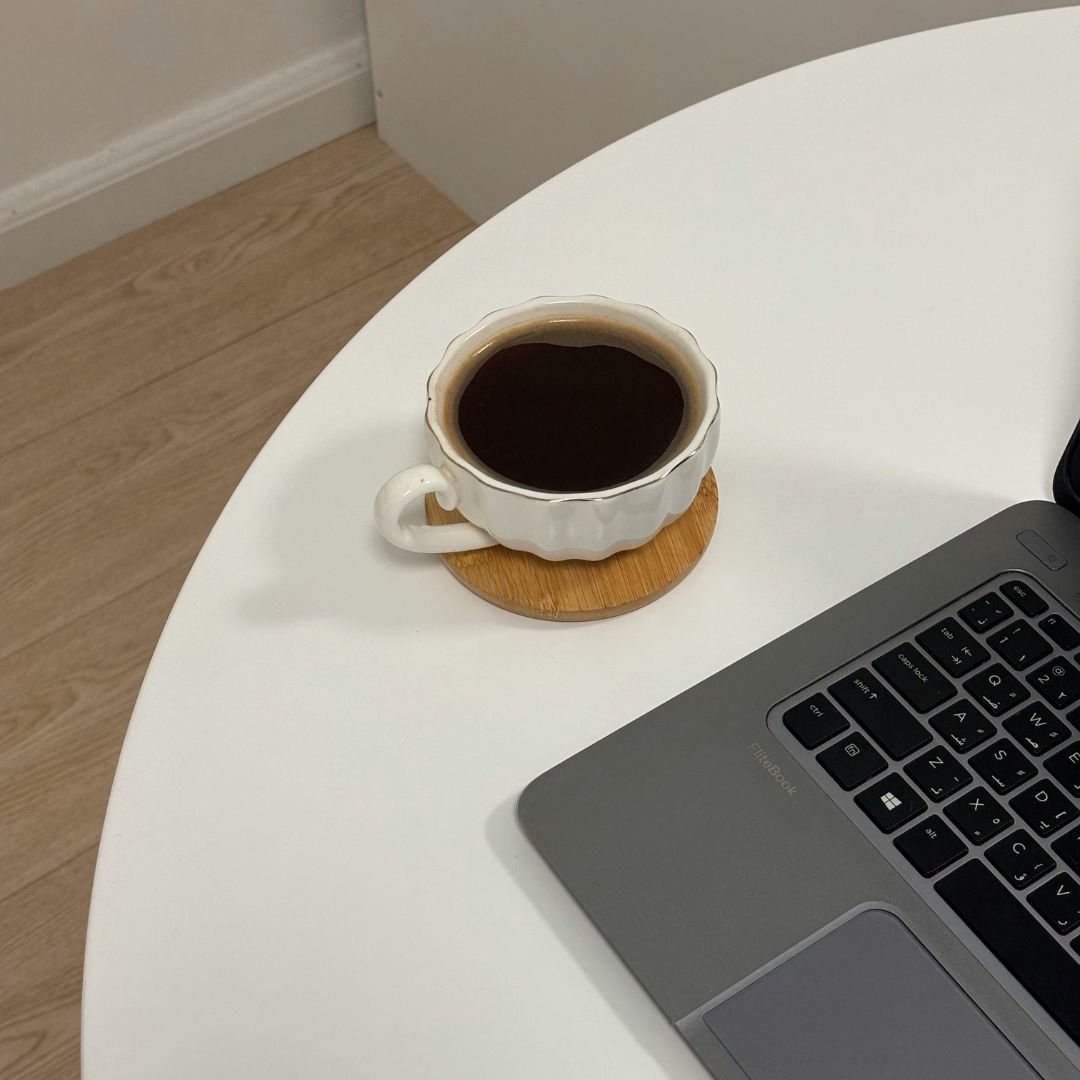 White circular table with a cup of coffee in a scalloped white mug and a silver laptop on top.