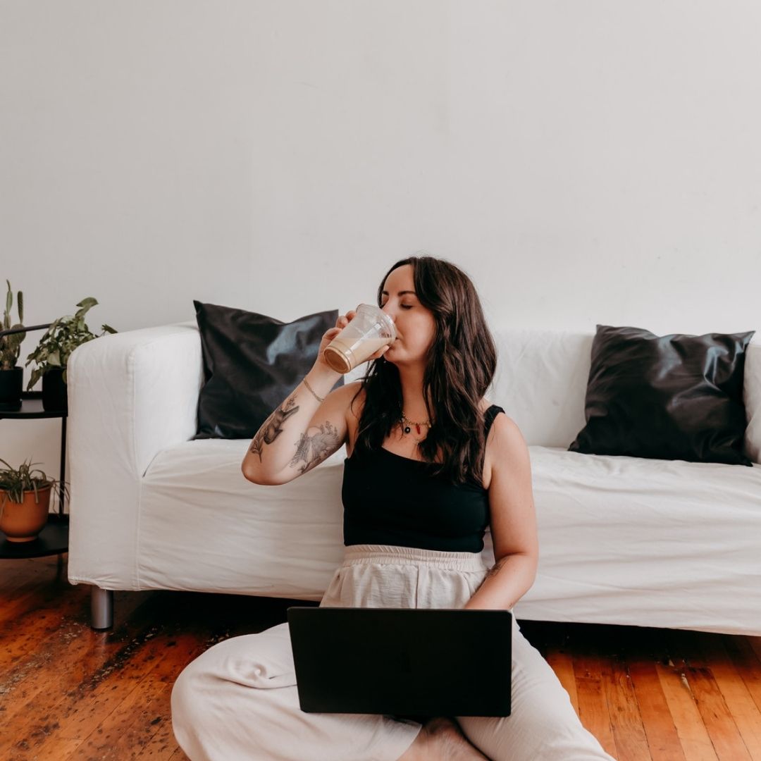 Delia, a brand strategist and SEO specialist, sitting on a wood floor in a sunlit studio in Baltimore. There's a white couch behind her, she has her laptop on her legs, and she is sipping an iced latte.