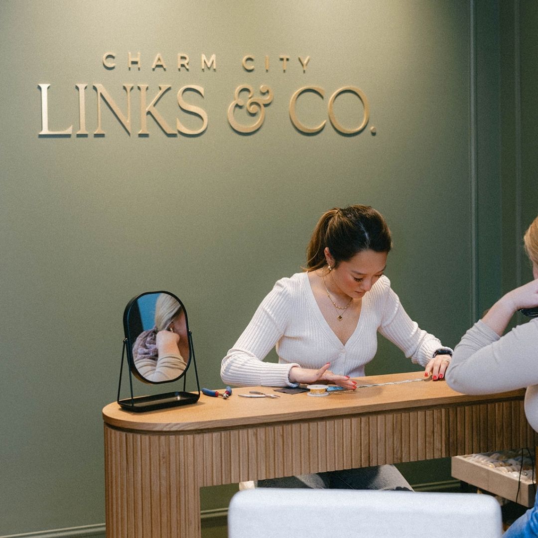 Young brunette permanent jeweler at Charm City Links in Catonsville measuring a 14k gold bracelet for a customer in their storefront. The wall is olive, they are sitting at a modern wood table, and there is a gold Charm City Links & Co. logo on the wall behind them.